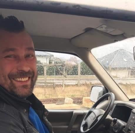 Uśmiechnięty mężczyzna z irokezem siedzi za kierownicą samochodu dostawczego, widok na domy w tle przez szybę.