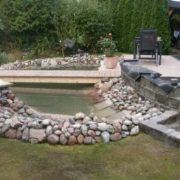 Oczko wodne z kamiennym brzegiem i drewnianym mostkiem, w tle widoczna altana i krzesło ogrodowe.