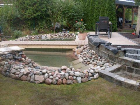 Oczko wodne z kamiennym brzegiem i drewnianym mostkiem, w tle widoczna altana i krzesło ogrodowe.