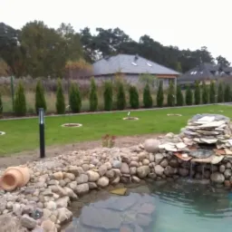 Ogród z trawnikiem, rzędem tuj, oczkiem wodnym z kaskadą z kamieni i terakotową donicą leżącą na brzegu.