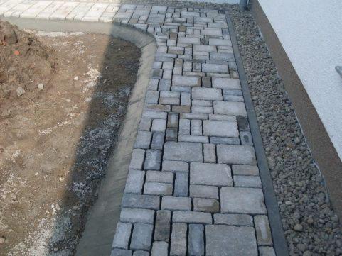 Fragment ścieżki z szarej kostki brukowej ułożonej w nieregularny wzór, z widoczną krawędzią z drobnego żwiru przy ścianie budynku i przygotowanym podłożem po drugiej stronie.