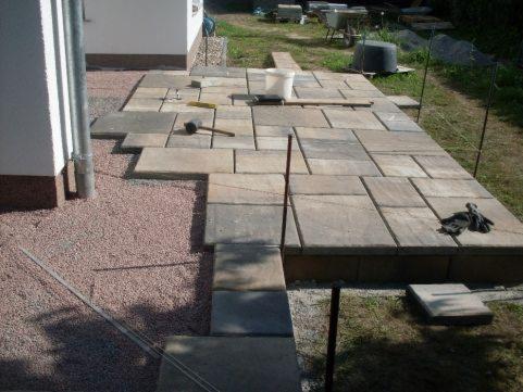 Układanie kostki brukowej na podwórku, widoczne narzędzia: gumowy młotek, poziomica, rękawice robocze, częściowo ułożona nawierzchnia z kostki w odcieniach szarości i beżu, obok wysypany żwir.