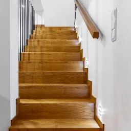Jasne, drewniane schody z minimalistyczną balustradą z czarnych prętów i drewnianą poręczą, oświetlone punktowymi lampami w ścianie, widok z dołu.