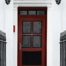 Czerwone drzwi wejściowe z czarnymi detalami i czterema szybami, osadzone w białej fasadzie z czarnymi, ozdobnymi lampami po bokach.