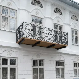 Elewacja budynku z białego kamienia z drewnianym balkonem z czarną, metalową balustradą o wzorze koncentrycznych półokręgów, widoczne okna z dekoracyjnymi nadprożami.