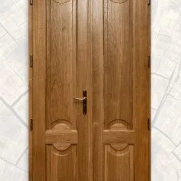 Dwu-skrzydłowe drzwi z jasnego drewna z ozdobnymi panelami i mosiężnymi zawiasami, widoczny detal faktury drewna i logo producenta w prawym dolnym rogu.