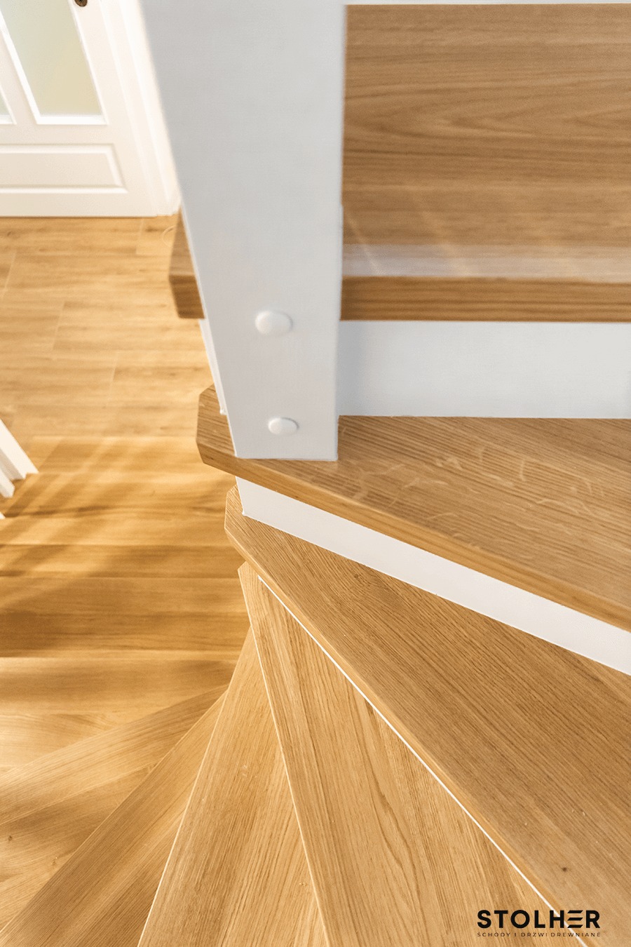 Drewniane schody spiralne z jasnego drewna dębowego, białe podstopnice i balustrada, widok z góry na zakręt schodów.