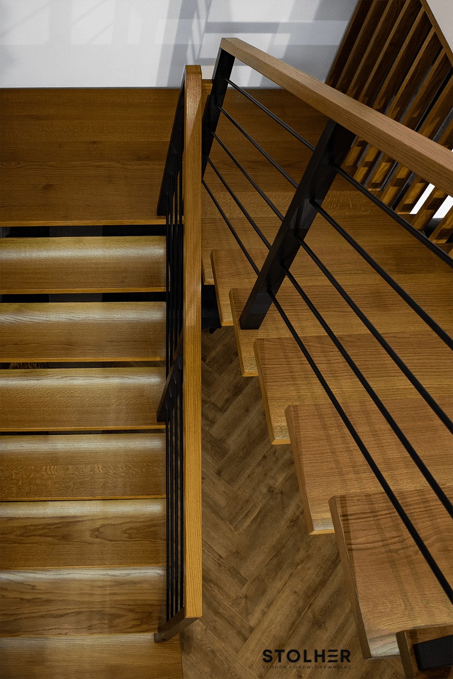 Drewniane schody z podświetleniem LED na stopniach, czarne metalowe poręcze, widok z góry na parkiet ułożony w jodełkę, logo firmy w rogu.