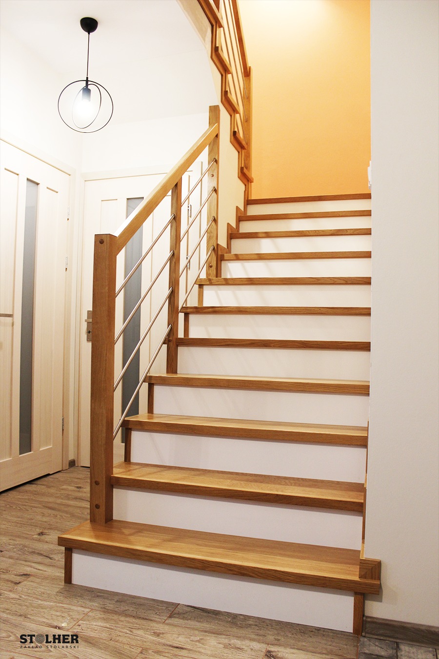 Drewniane schody z białymi podstopnicami i metalowymi poręczami, oświetlone wiszącą lampą z okrągłym kloszem, widoczne drzwi w tle.