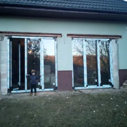 Nowe, dwuskrzydłowe okna balkonowe z białymi ramami marki VEKA, jeszcze z folią ochronną, zamontowane w surowym otworze ściany budynku; dziecko w kurtce stoi przed nimi z uniesionymi rękami.