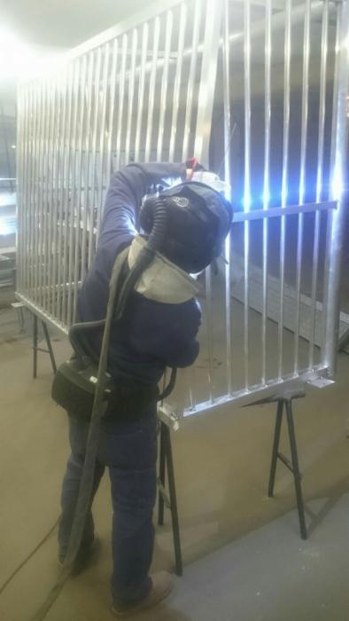 Pracownik w kombinezonie ochronnym spawa metalową konstrukcję z pionowych prętów na warsztacie, widoczne iskry i sprzęt spawalniczy.