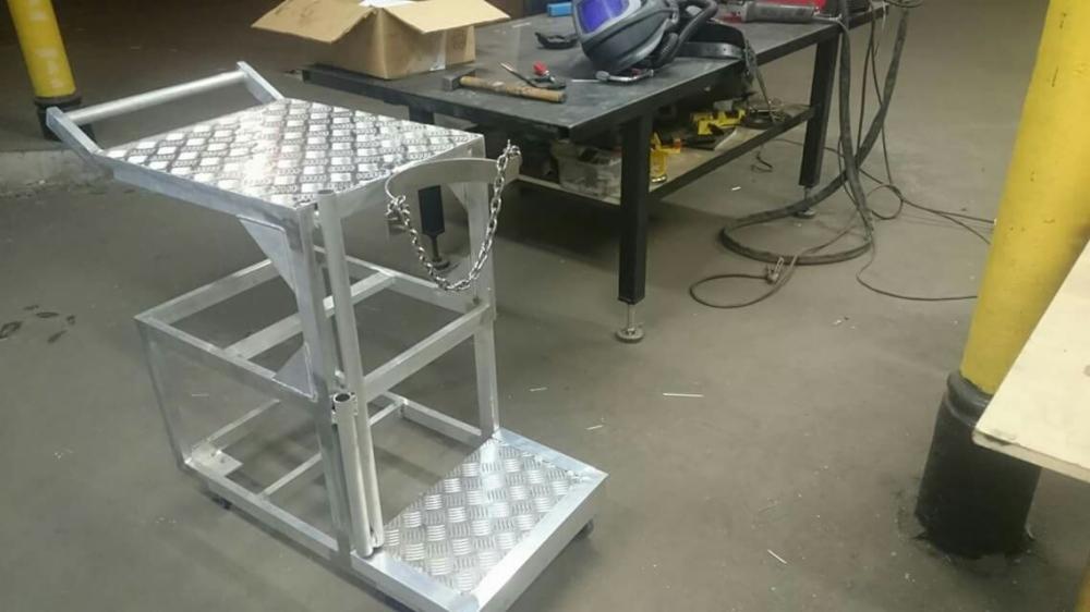 Metalowa konstrukcja w warsztacie, prawdopodobnie wózek lub podest roboczy, z blachą ryflowaną na platformach, obok stoi stół warsztatowy z narzędziami i hełmem spawalniczym.