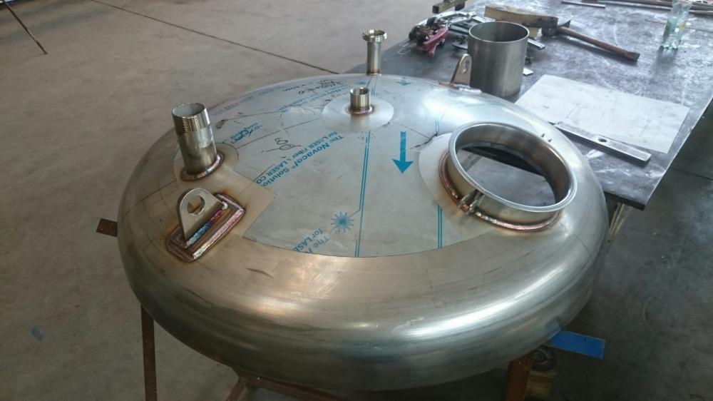 Zbliżenie na metalowy zbiornik ciśnieniowy z przyspawanymi króćcami i uchwytem, widoczne ślady po folii ochronnej i niebieska strzałka na powierzchni.