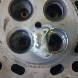 Głowica silnika samochodowego po naprawie, z widocznym świeżym spawem wypełniającym obszar między gniazdami zaworowymi.