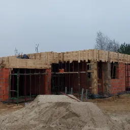 Wznoszenie ścian parterowego domu z czerwonej cegły, widoczne szalunki i zbrojenia stropu, w tle las.