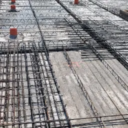 Zbrojenie stropu domu jednorodzinnego z widocznymi rurami wentylacyjnymi, przygotowane do zalania betonem, na tle budynku mieszkalnego i zieleni.