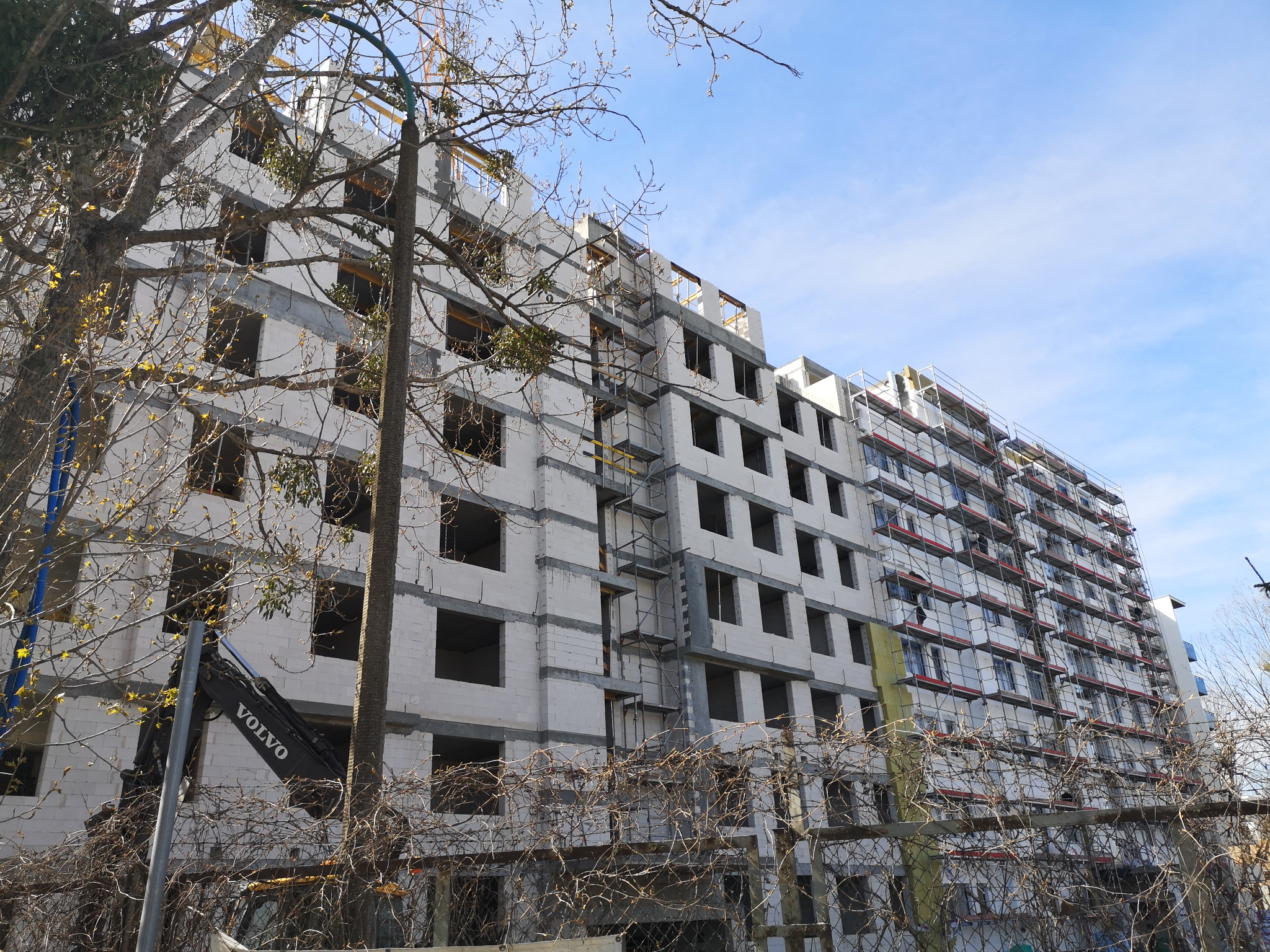 Wielopiętrowy budynek w stanie surowym, z białych bloczków, z widocznymi rusztowaniami i żurawiem Volvo w tle, częściowo przysłonięty gałęziami drzew