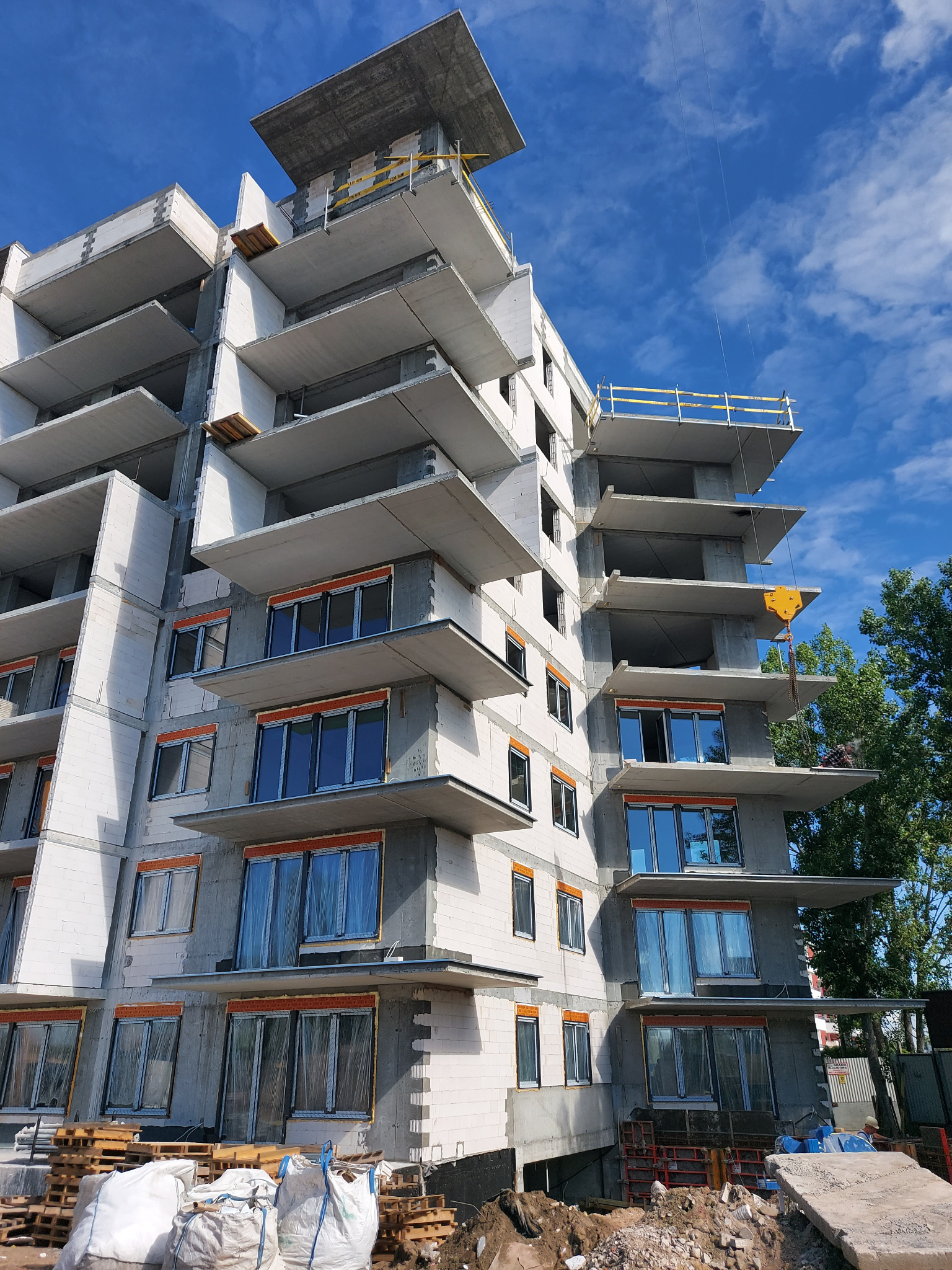 Wielopiętrowy budynek w trakcie budowy, z widocznymi białymi bloczkami i wystającymi balkonami, okna zabezpieczone folią, żółty dźwig w tle, błękitne niebo.