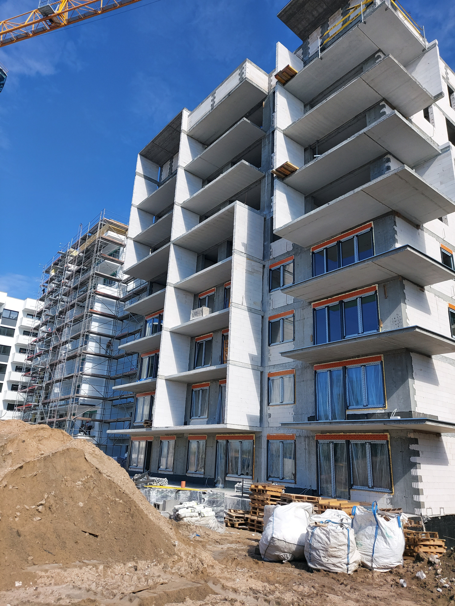 Wznoszony budynek wielorodzinny z betonu komórkowego z widocznymi balkonami, oknami zabezpieczonymi folią, rusztowaniami i stertą ziemi na pierwszym planie.