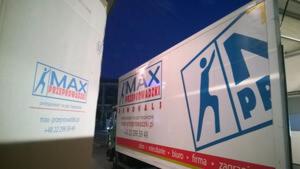 Dwa białe samochody dostawcze z logo firmy transportowej MAX Przeprowadzki, widoczne z boku, z niebieskim logotypem i danymi kontaktowymi, sfotografowane wieczorem.