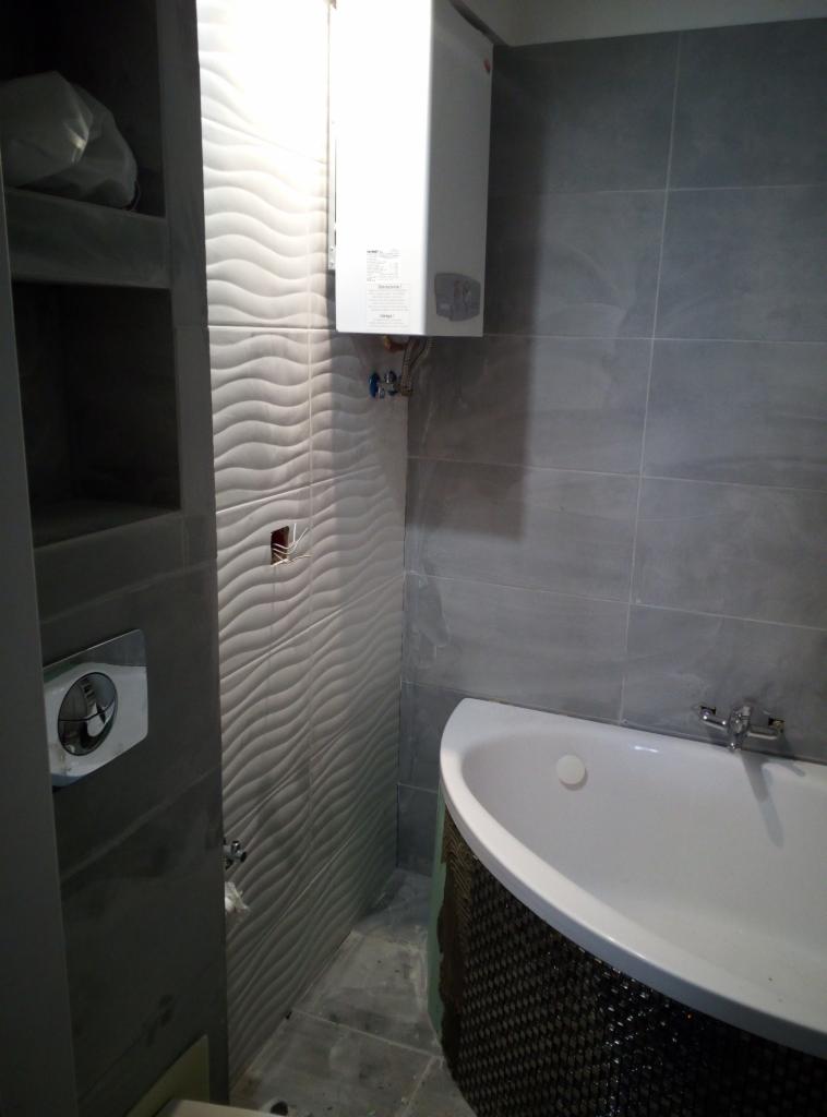 Szara łazienka z wanną narożną, grzejnikiem elektrycznym i falistymi płytkami na ścianie, widoczne niedokończone wykończenie otworu na gniazdko elektryczne.