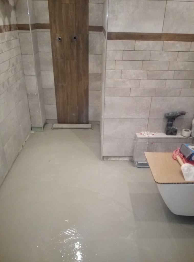 Wyrównana wylewka samopoziomująca w łazience z częściowo położonymi płytkami na ścianach, widoczna kabina prysznicowa z drewnopodobnymi panelami i narzędzia glazurnicze.