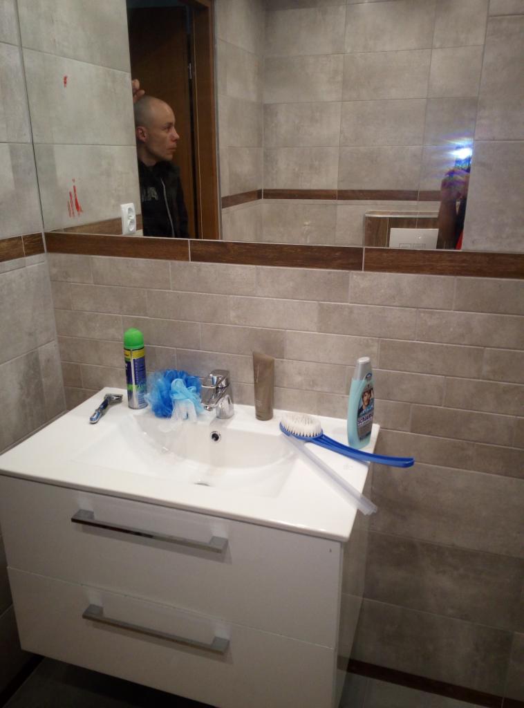 Umywalka z białymi szafkami w łazience wyłożonej szarymi płytkami, widoczne kosmetyki i odbicie osoby w lustrze.