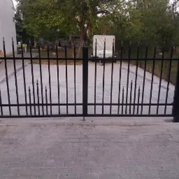 Czarna, metalowa brama wjazdowa z dekoracyjnymi grotami, widok na podjazd wyłożony kostką brukową, w tle drzewo i przyczepa.