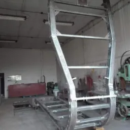 Konstrukcja metalowa spawana w warsztacie, przypominająca fragment bramy przemysłowej lub element większej konstrukcji, surowe wykończenie, widoczne ślady spawania, podparta cegłami.