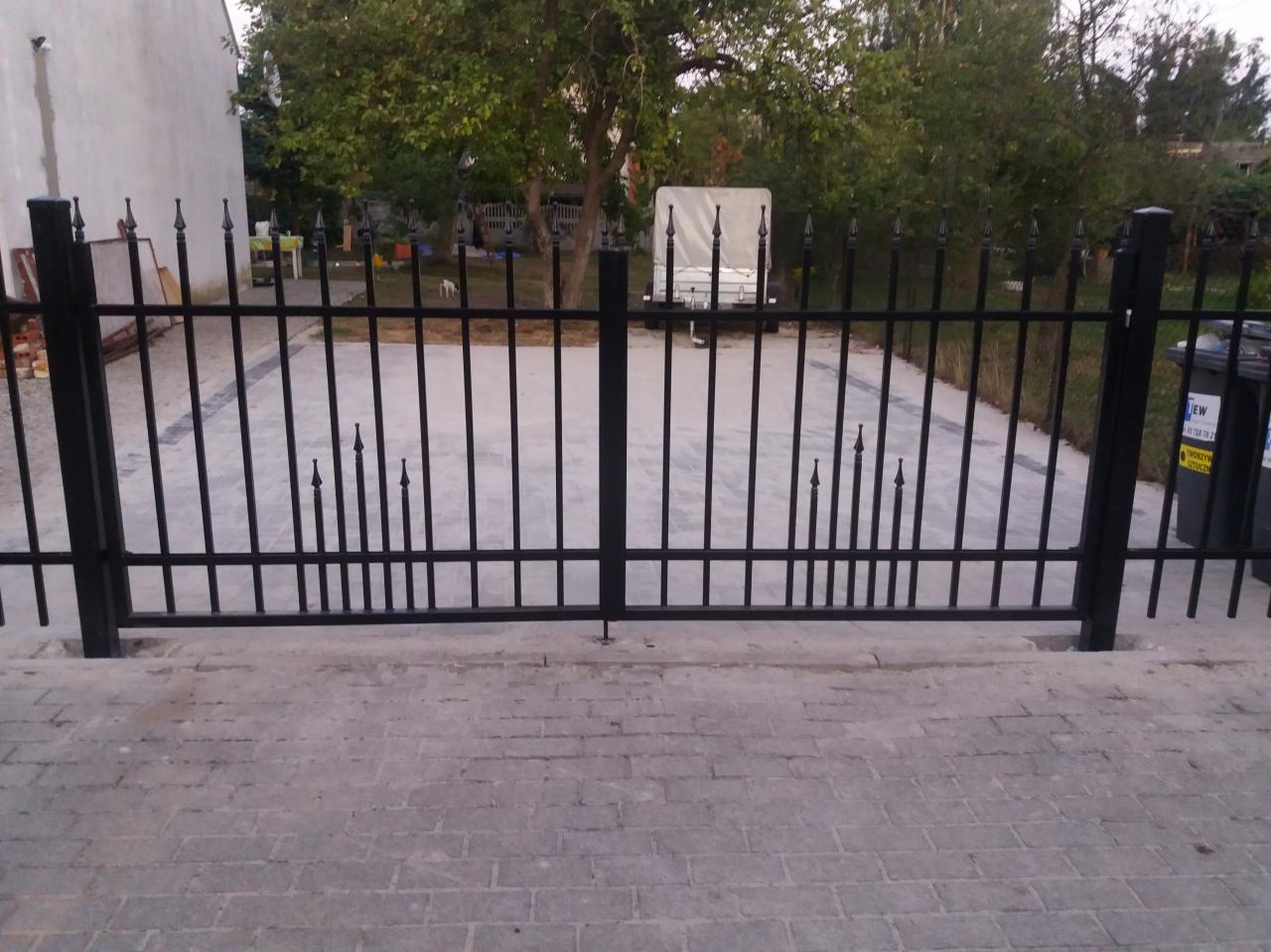Czarna, metalowa brama wjazdowa z dekoracyjnymi grotami, widok na podjazd wyłożony kostką brukową, w tle drzewo i przyczepa.