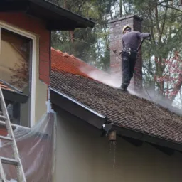 Pracownik w kasku czyści dach z mchu i zabrudzeń za pomocą myjki ciśnieniowej, widoczny komin, drabina oparta o ścianę budynku.