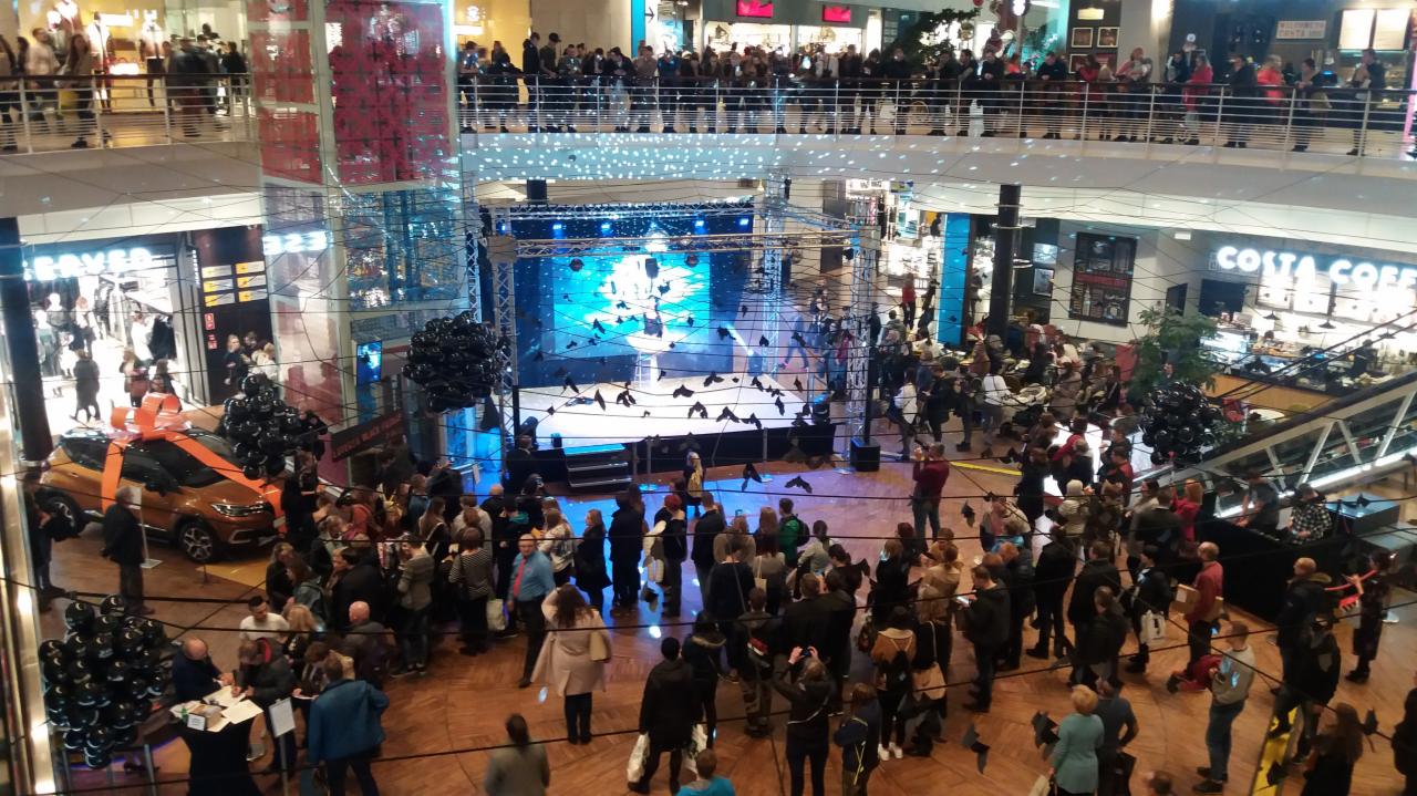 Noc Zakupów Black Friday w Manufakturze w Łodzi: scena z niebieskim oświetleniem, tłum ludzi na parterze i balkonach, samochód z pomarańczową kokardą i czarne balony.