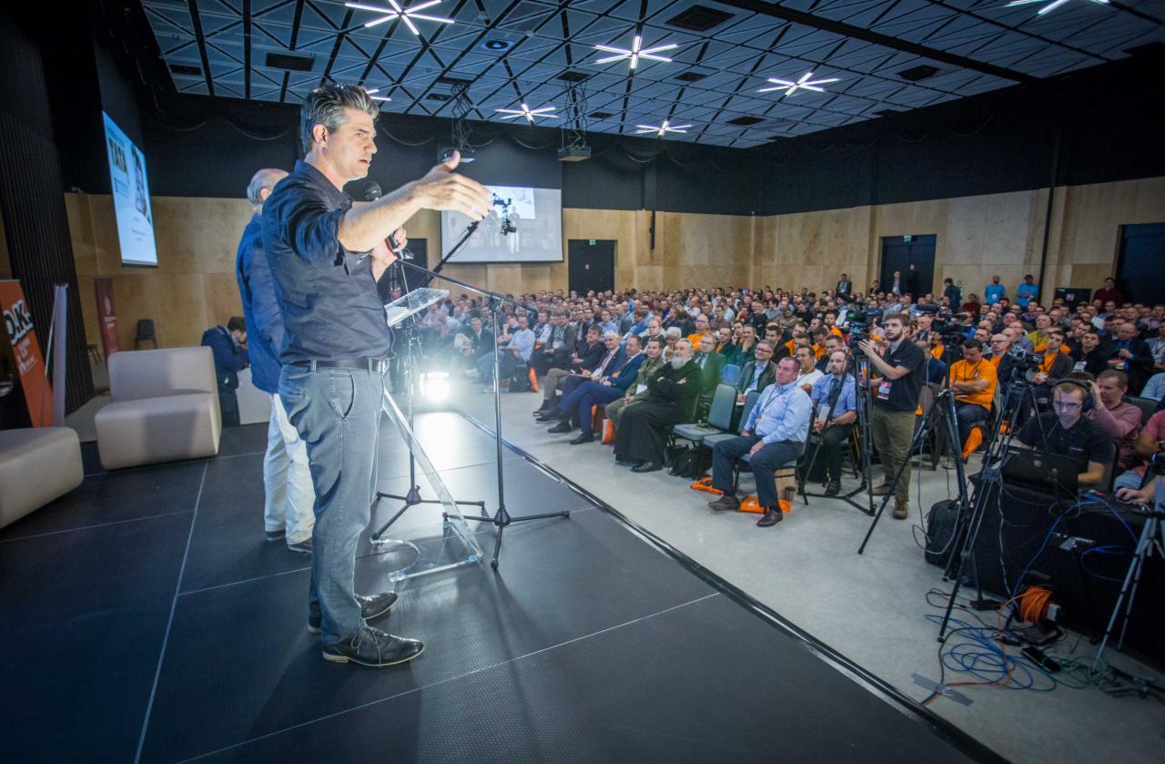 Prelegent z mikrofonem na scenie podczas konferencji, za nim widoczny ekran i publiczność w sali konferencyjnej z charakterystycznym oświetleniem sufitowym.