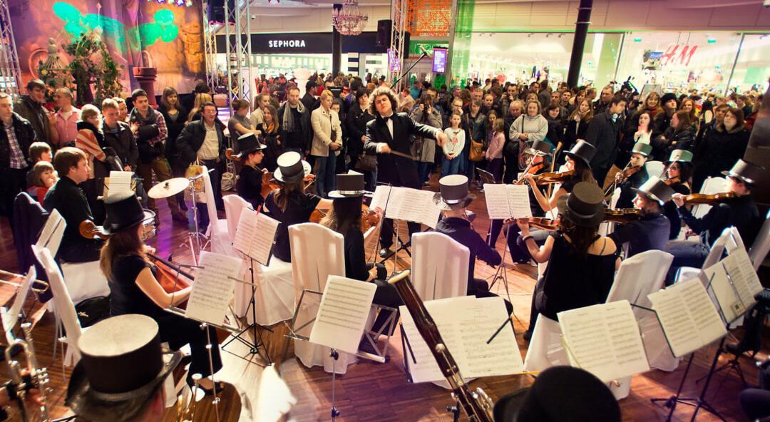 Orkiestra w cylindrach gra koncert w centrum handlowym Manufaktura Łódź podczas Black Friday, tłum widzów obserwuje występ na tle sklepów Sephora i H&M.