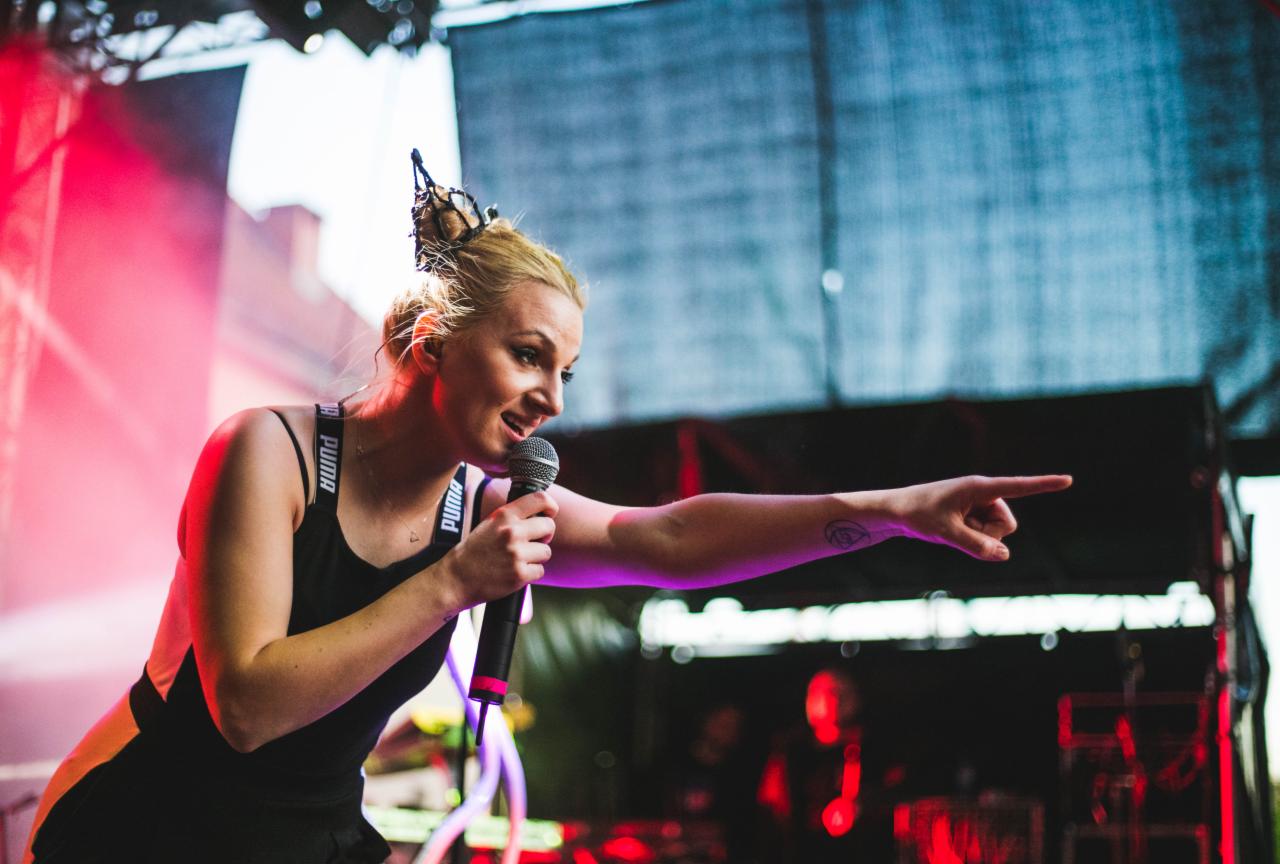 Wokalistka z mikrofonem na scenie podczas koncertu, wskazująca palcem w dal, z ozdobą we włosach w kształcie motyla i tatuażem na ramieniu.