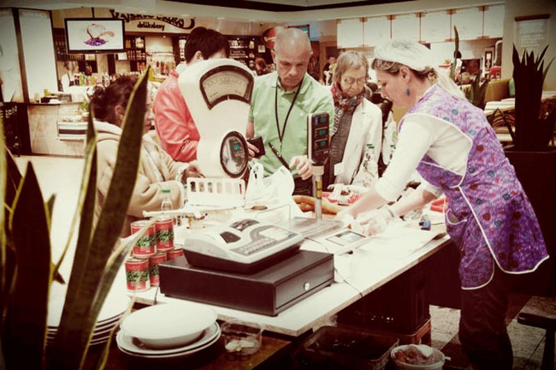 Obsługa klienta w sklepie spożywczym: ekspedientka w fioletowym fartuchu waży wędliny na wadze elektronicznej dla kolejki klientów, w tle widoczne półki z produktami.