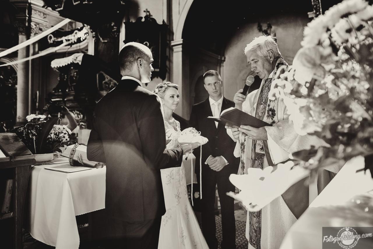 Czarno-białe ujęcie z ceremonii ślubnej w kościele: panna młoda z bukietem, pan młody tyłem, urzędnik z mikrofonem i księgą, druh w tle.