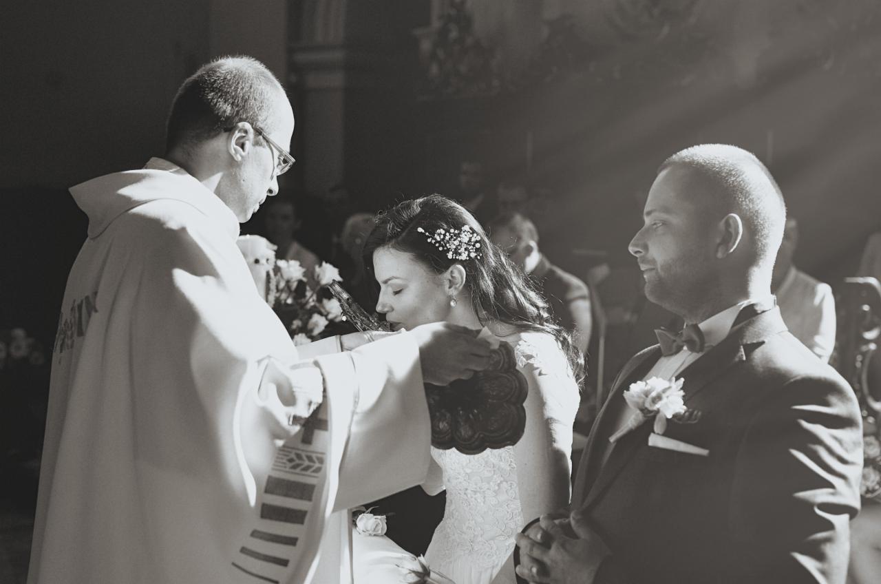 Czarno-białe zdjęcie z ceremonii ślubnej w kościele: panna młoda całuje krucyfiks podawany przez księdza, obok stoi pan młody w garniturze z muszką, w tle widoczni goście.