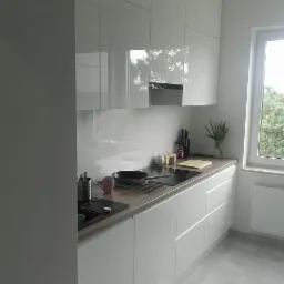 Nowoczesna, minimalistyczna kuchnia z białymi, lakierowanymi frontami szafek i blatem imitującym drewno. Na blacie widoczny czarny zlew, płyta indukcyjna, naczynia i akcesoria kuchenne. W tle okno...