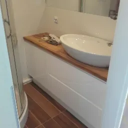 Biała umywalka nablatowa na drewnianym blacie w łazience z brązowymi płytkami podłogowymi, widoczne narzędzia montażowe na blacie.