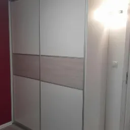 Dwudrzwiowa szafa przesuwna z białymi frontami i poziomym pasem w kolorze jasnego drewna, srebrne obramowanie, widoczny fragment bordowej ściany w tle.