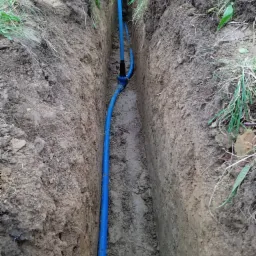 Wąski wykop w ziemi z ułożoną niebieską rurą instalacyjną, widoczne ślady po wykopie i fragmenty darni na krawędziach.