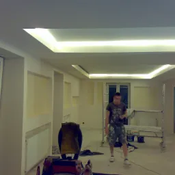 Wnętrze w trakcie remontu z podwieszonym sufitem z oświetleniem LED, widoczny pracownik w ubraniu roboczym, rusztowanie oraz narzędzia malarskie.