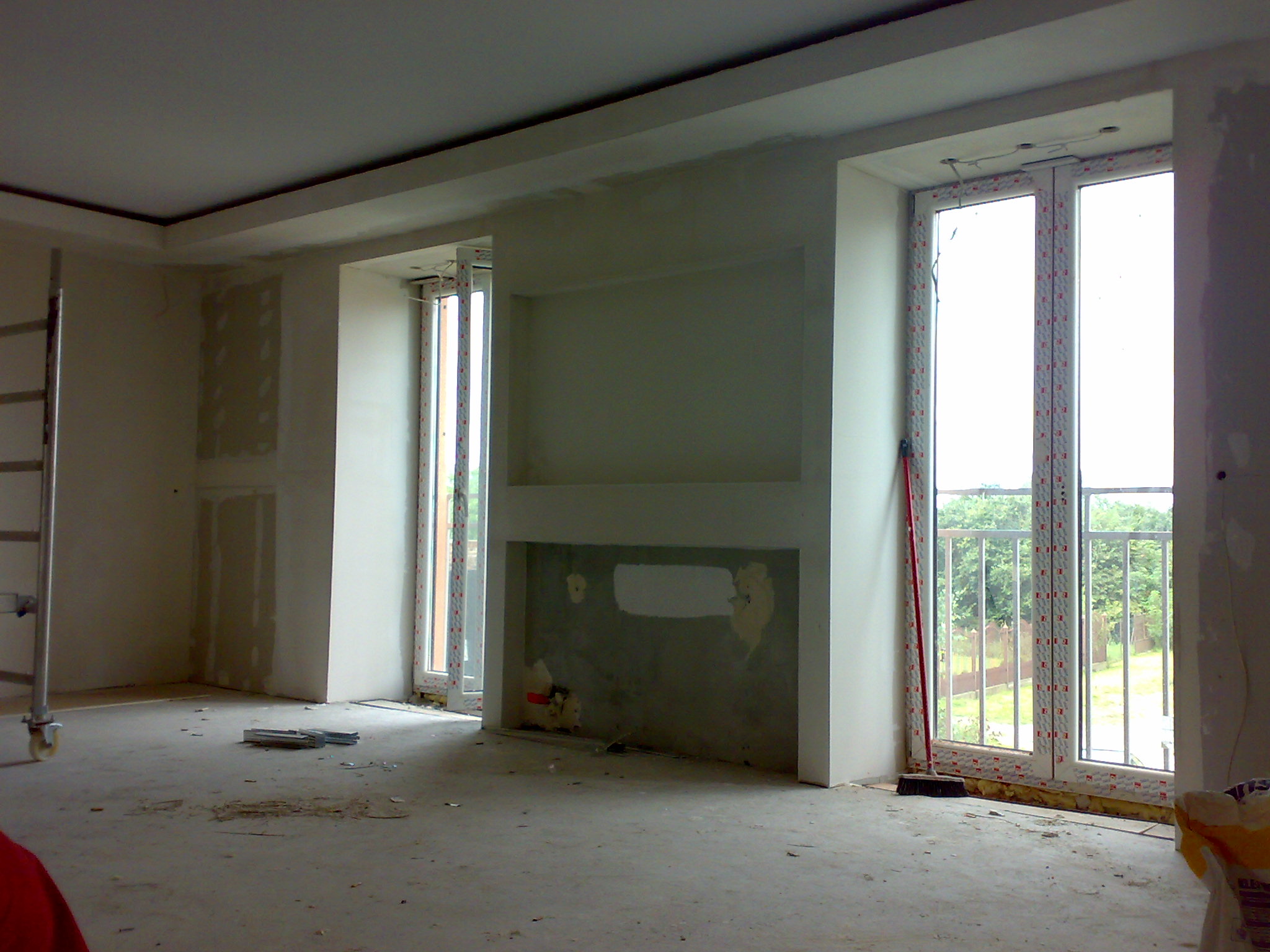 Pokój w trakcie remontu: szpachlowane ściany, surowy beton, okna PCV z widokiem na zieleń, drabina aluminiowa w rogu.