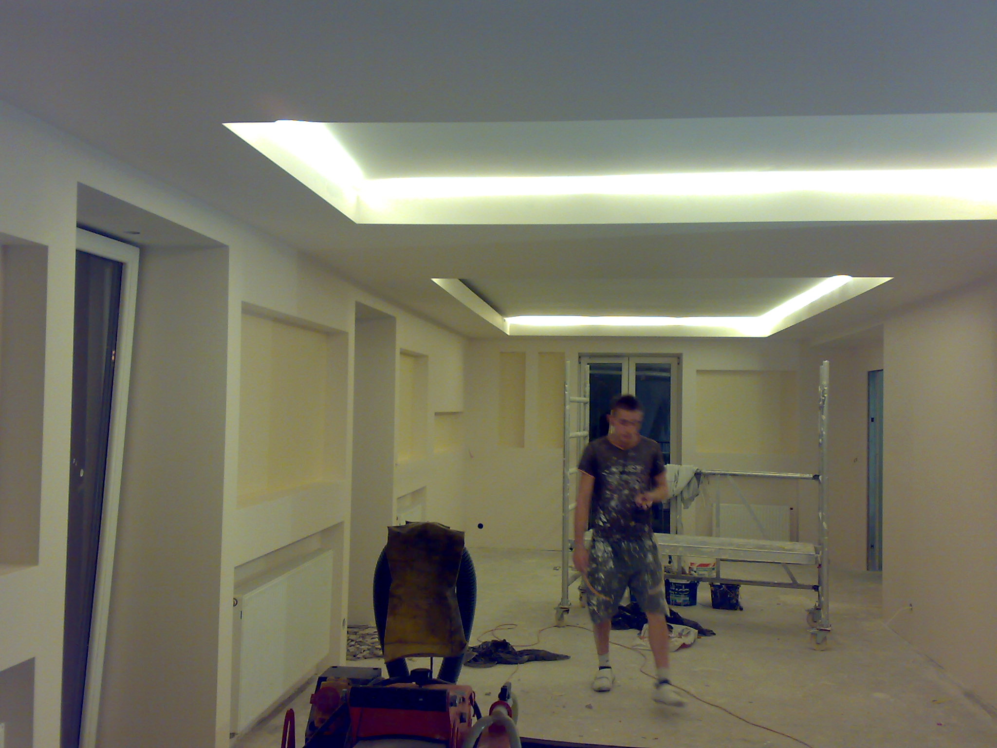 Wnętrze w trakcie remontu z podwieszonym sufitem z oświetleniem LED, widoczny pracownik w ubraniu roboczym, rusztowanie oraz narzędzia malarskie.