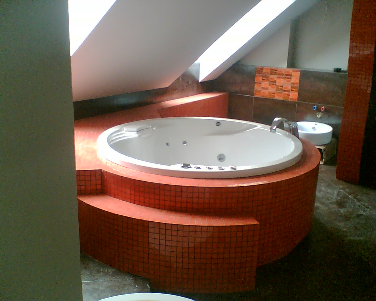 Okrągła wanna z hydromasażem obudowana mozaiką w odcieniach pomarańczy w łazience na poddaszu ze skośnym oknem dachowym.
