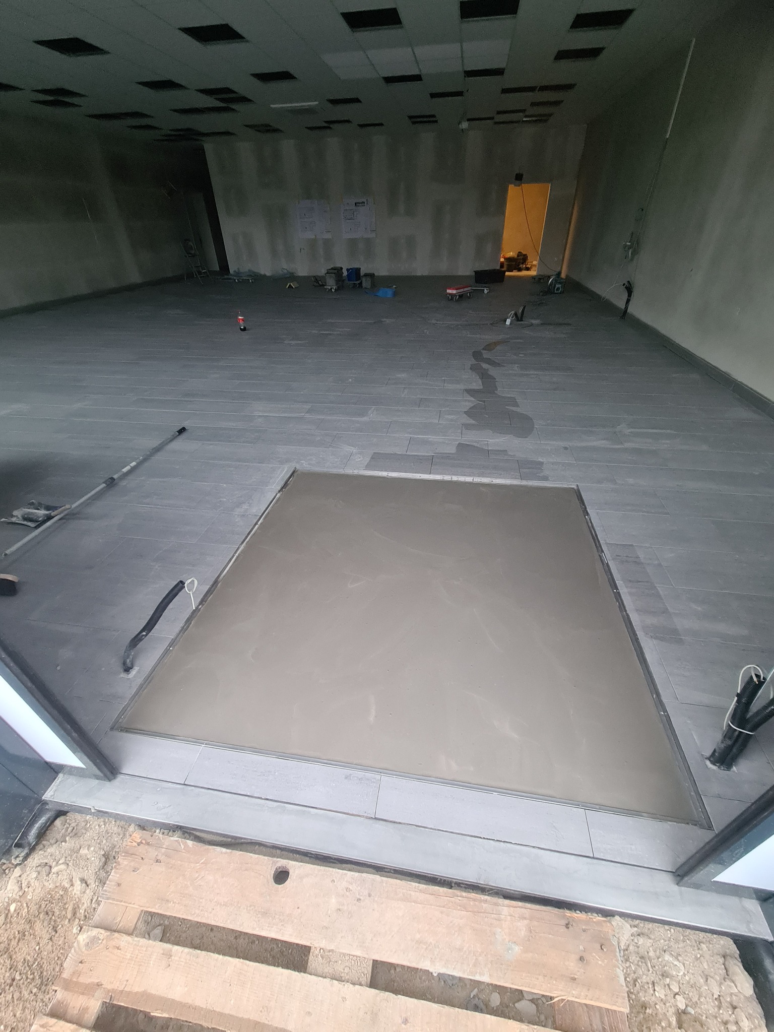 Wnętrze w trakcie prac glazurniczych: szare płytki podłogowe, wylewka samopoziomująca w kwadratowej wnęce przy wejściu, widoczne narzędzia i materiały budowlane w tle, sufit z rastrem oświetleniowym.