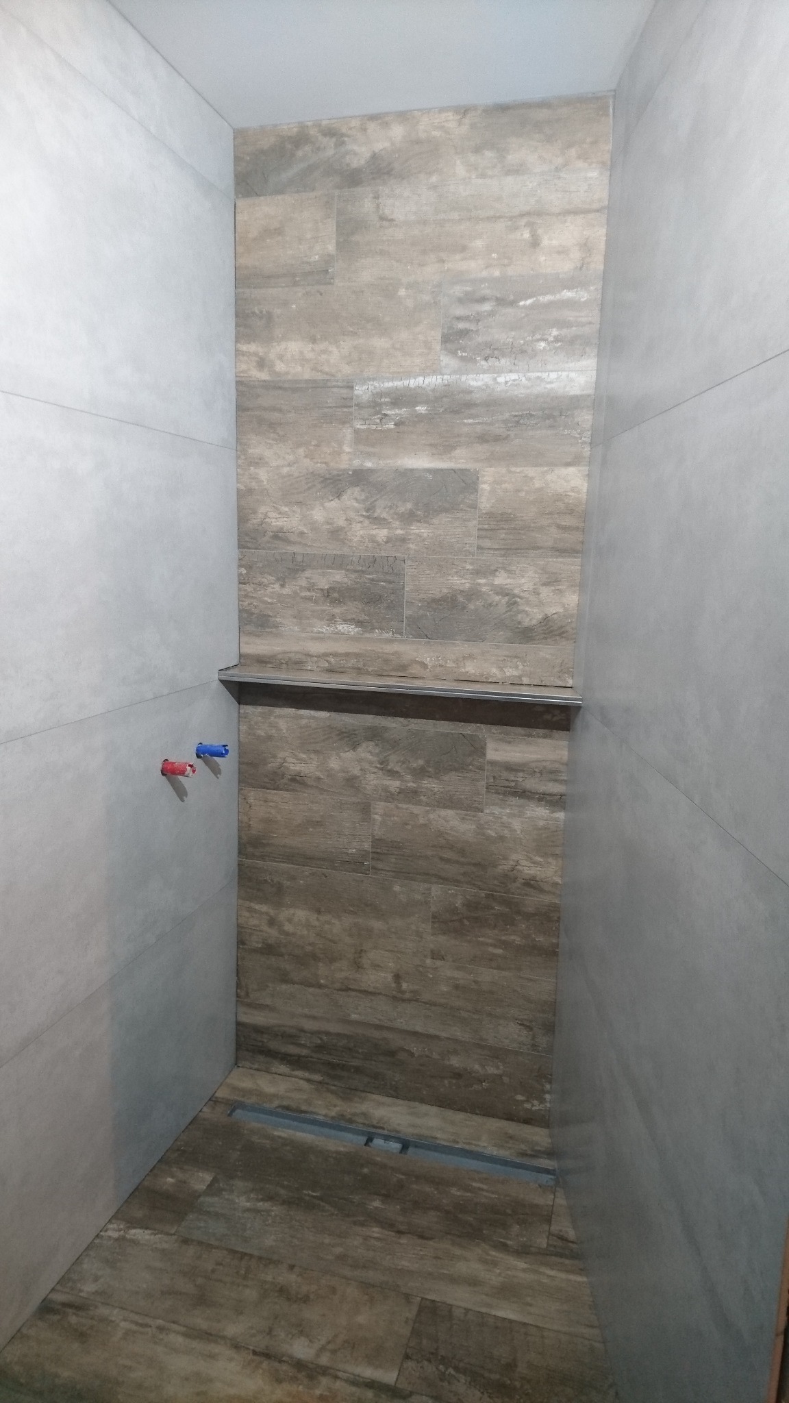 Wykończona kabina prysznicowa z odpływem liniowym i półką, wyłożona płytkami imitującymi drewno na ścianie akcentującej i szarymi płytkami na pozostałych ścianach.