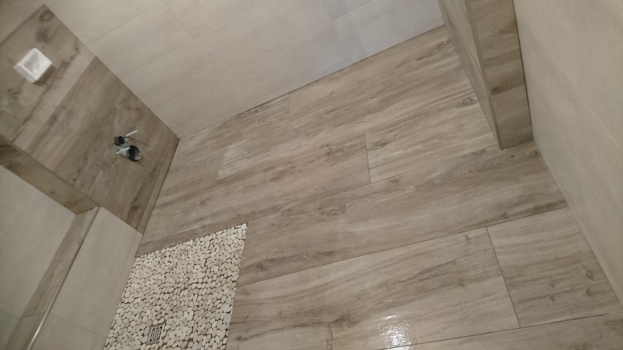 Wykończona łazienka z podłogą imitującą drewno i dekoracyjnym fragmentem z białych kamieni przy odpływie.