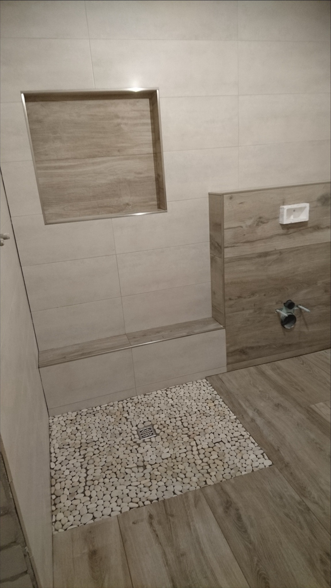 Beżowa łazienka z kabiną prysznicową wyłożoną białymi kamieniami, odpływ liniowy, wnęka w ścianie z drewnopodobną okładziną, zabudowa podtynkowego WC z okładziną drewnopodobną.